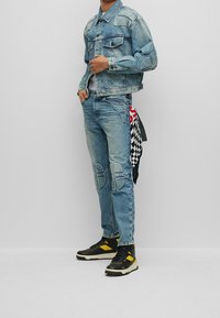 Ljusblå denimjacka med patchdetaljer, tillsammans med matchande jeans med sidofickor och en rutmönstrad bandana knuten i midjan. Svarta sneakers med gula accenter.