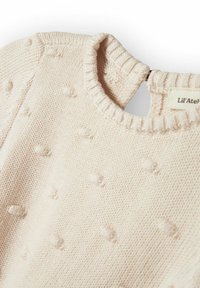 Lys beige strikket genser med rund hals, med et strukturert boblemønster og ribbet krage. Etikett synlig bak.