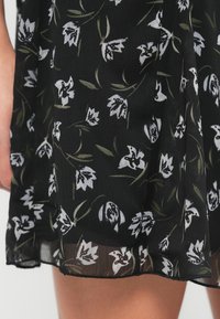 Μαύρο διαφανές ύφασμα με λευκό και πράσινο λουλουδένιο μοτίβο σε ένα κοντό φόρεμα, που καλύπτει μερικώς το πάνω μέρος του μηρού και το χέρι ενός ατόμου.