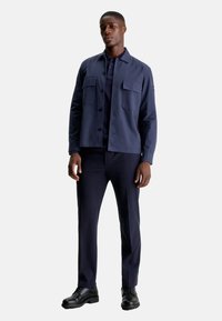 Giacca in cotone blu navy con due tasche anteriori, chiusura a bottone e vestibilità slim, abbinata a pantaloni coordinati e scarpe nere.