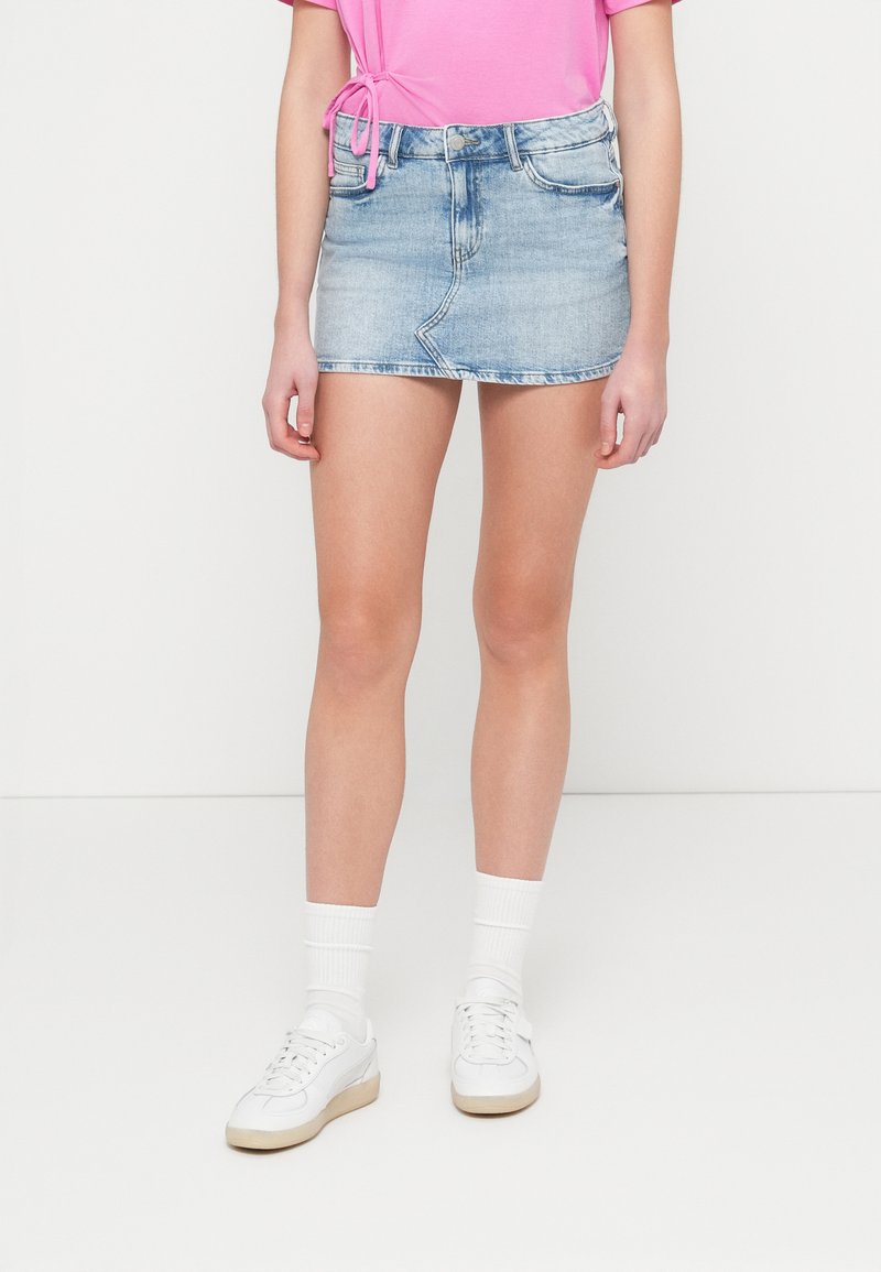 Τζιν μίνι φούστα σε ανοιχτό μπλε χρώμα, με εφαρμοστή μέση και ξέφτια στο τελείωμα, συνδυασμένη με ροζ τοπ και λευκά αθλητικά παπούτσια.