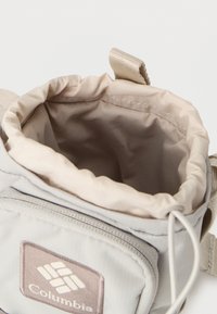 Φωτεινό γκρι υφασμάτινο πουγκί με κορδόνι στο πάνω μέρος, με απαλό εσωτερικό επένδυση και μικρό λογότυπο Columbia σε καφέ ετικέτα.