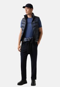 Mand iført sort baseballcap, navyblå puffervest over blå t-shirt, mønstrede sweater bundet om taljen, navybukser og sorte støvler.