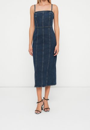 Femme portant une robe midi en jean bleu foncé ajustée, sans manches, avec des fines bretelles et des sandales noires à talons hauts avec brides à la cheville.