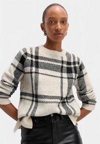 Crème trui met zwart geruit patroon, ronde hals en geribbelde manchetten. Draagt zwart leren broek met zijopeningen aan de zoom.