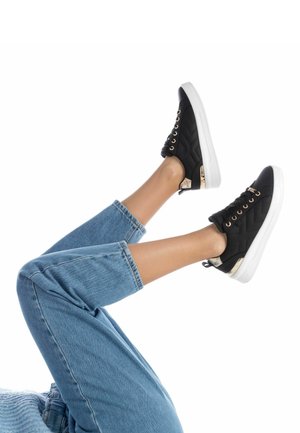 Zapatillas acolchadas negras con ojales y acentos dorados, combinadas con pantalones vaqueros de corte ligero azul claro, exhibidas sobre piernas elevadas contra un fondo blanco.