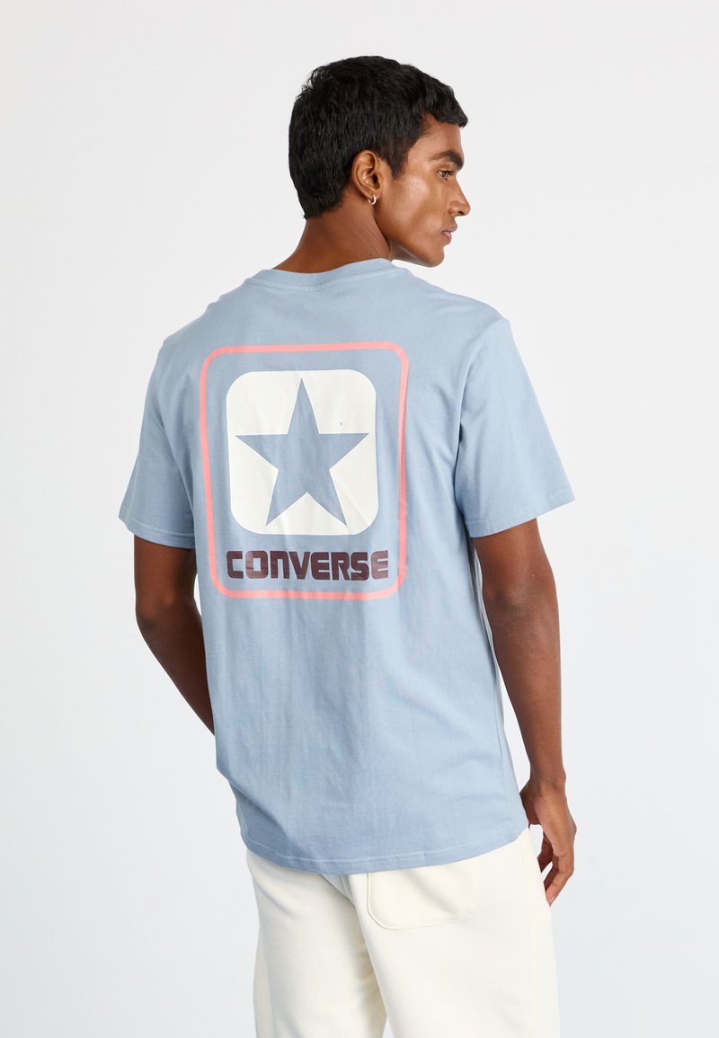 Lyseblå bomulds T-shirt med et stort hvidt stjernegrafik og "CONVERSE" tekst i brun, med en afslappet pasform og korte ærmer.