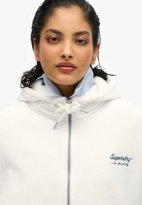 Witte zip-up hoodie met een zachte structuur, voorzien van een trekkoordcapuchon en een blauw geborduurd logo op de borst. Lichte blauwe kraag zichtbaar.