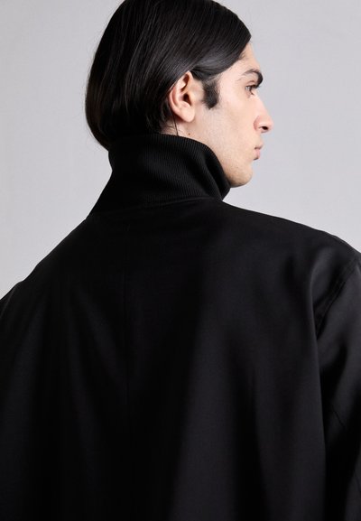 Black outerwear with a high collar, featuring a smooth texture and loose fit, shown from the back. Hair is long and straight.