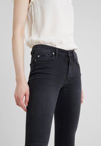 Jeans ajustados negros con una textura suave, que cuentan con un bolsillo delantero, accesorios metálicos y un sutil detalle de logo en el bolsillo.