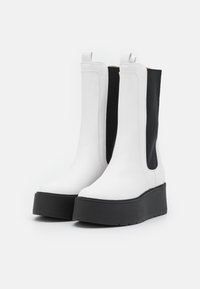 Botas brancas até ao tornozelo com painéis laterais em tecido preto, um upper em couro liso e uma solinha grossa preta. Apresentam puxadores para um calçar fácil.