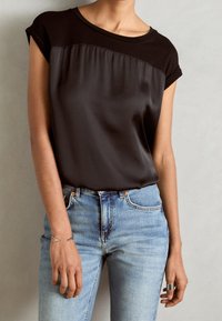 Osoba w czarnej satynowej bluzce z krótkim rękawem włożonej w jasnoniebieskie dżinsy z wysokim stanem, z srebrną bransoletką i pierścionkiem na lewej i prawej ręce.