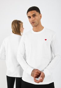 Λευκό μακρυμάνικο μπλουζάκι με κόκκινο έμβλημα καρδιάς στο στήθος. Μαλακό ύφασμα βαμβακιού με στρογγυλή λαιμόκοψη και εφαρμοστά μανίκια.
