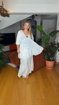 White tiered dress with long sleeves, textured fabric, and a V-neck. Worn with sandals, displayed in a room with a green plant and sofa.