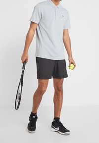 Grå polo skjorta med en liten grön logotyp, kombinerad med svarta shorts. Håller en tennisracket i ena handen och en tennisboll i den andra. Svarta skor.