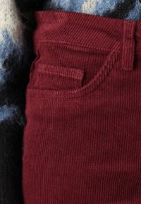 Burgundské kordurové látky ukazující detail kapsy kalhot, s texturovanými žebry a kontrastním prošíváním.
