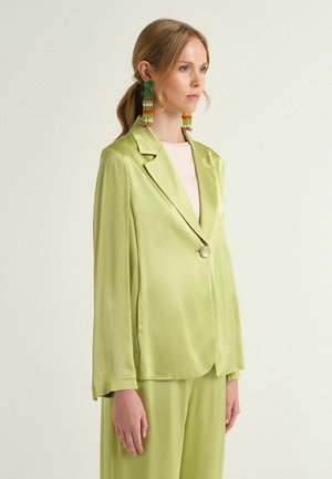 Frau in grünem Satin-Blazer mit großem Knopf und passenden weiten Hosen, trägt mehrfarbige, mit Perlen verzierte Quastenohrringe, schaut nach rechts.