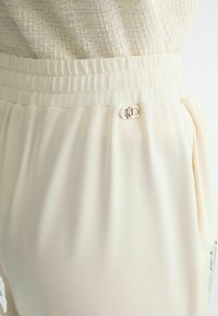 Gros plan sur un pantalon crème avec ceinture élastique et un petit logo métallique à double anneau sur la poche côté droit.