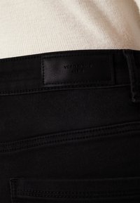 Crne traperice od denima imaju teksturiranu oznaku na pojasu označenu "VERO MODA." Jednostavno šivanje duž džepova i pojasa je vidljivo.