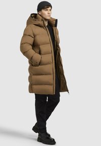 Brun oppustet frakke med hætte, kviltet design, for- og sidelommer samt lynlås, båret over en sort top og parret med sorte bukser og støvler.
