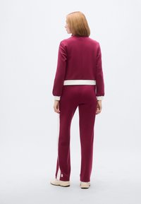 Μπορντό φόρμα με κοντό μπουφάν και μακριά παντελόνια. Λευκές λεπτομέρειες σε μανσέτες και πλαϊνές ρίγες. Λείο ύφασμα με καθαρό, εφαρμοστό σχέδιο.