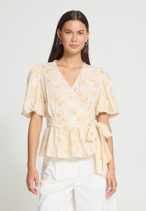 Femme portant un chemisier cache-cœur jaune clair à motifs floraux, avec des manches bouffantes et une taille nouée, associé à un pantalon blanc, debout devant un fond uni.