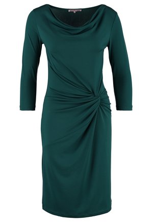 Kvinde iført en knælænge kjole i mørkegrøn med indsnørret talje, sorte gennemsigtige strømpebukser, sorte højhælede sko og en sort clutchtaske i hånden.