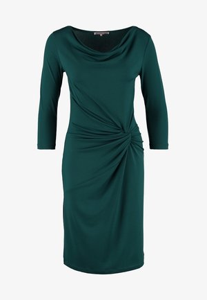 Kvinde iført en knælænge kjole i mørkegrøn med indsnørret talje, sorte gennemsigtige strømpebukser, sorte højhælede sko og en sort clutchtaske i hånden.