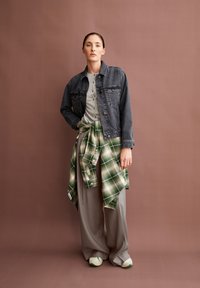 Frau steht vor einem braunen Hintergrund, trägt eine schwarze Jeansjacke, ein graues Hemd mit Knöpfen, ein grün kariertes Hemd, das um die Taille gebunden ist, weite graue Hose und Sneaker.