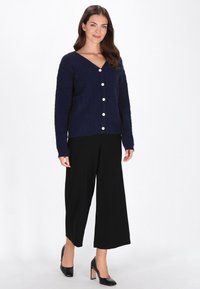 Marineblauwe gebreide cardigan met een V-hals, voorzien van een textuurpatroon en grote witte knopen, gecombineerd met zwarte wijde broek en zwarte hakken.