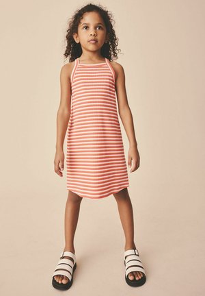 Ung pige med krøllet hår iført en ærmeløs orange og hvid stribet kjole og hvide sandaler, stående foran en beige baggrund.
