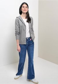Cardigan à capuche gris, t-shirt blanc, jeans évasés bleus, baskets blanches avec des accents clairs, debout devant un fond clair.