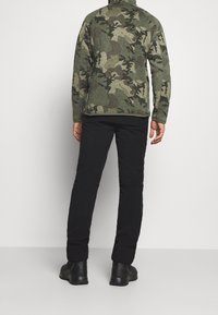 Veste en polaire camouflage vert avec col montant, portée avec un pantalon noir et des chaussures noires, vue de dos. Tissu et motif texturés visibles.