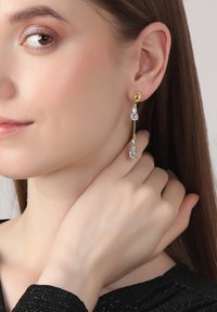 Goldfarbene Tropfenohrringe mit drei klaren Edelsteinen an einer zarten Kette, die unter dem Ohrläppchen hängen. Glatte und polierte Oberfläche.