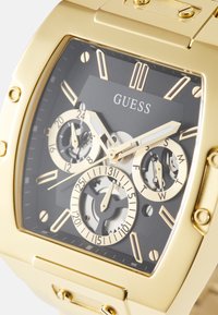 Montre carrée dorée avec cadran noir, trois sous-cadrans et accents en acier inoxydable. Comprend des marqueurs d'heure et des fonctions jour/date.