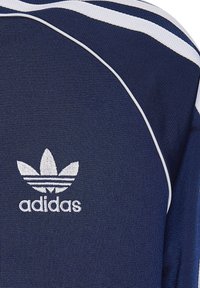 Sportovní tričko tmavě modré barvy s bílými linkami. Má texturovanou tkaninu a vyšívané logo Adidas se třemi listovými pruhy.