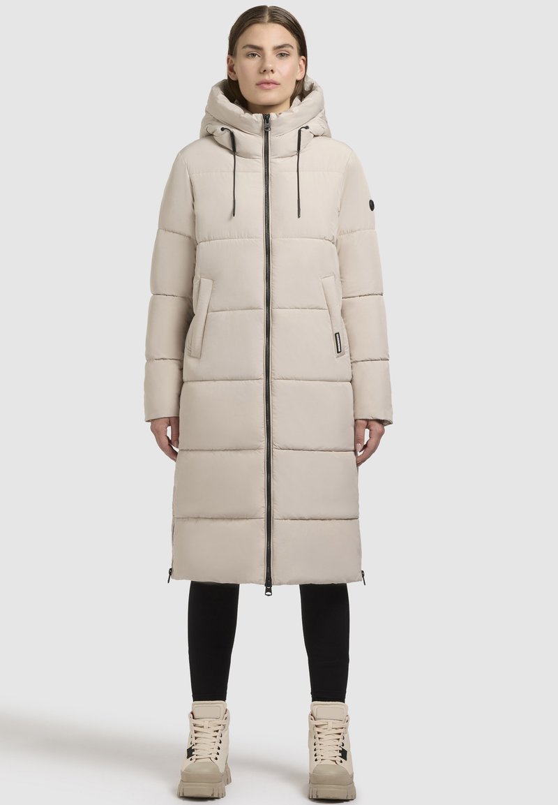 Hellbeige Pufferjacke mit Kapuze, vertikaler Steppung, Frontreißverschluss, zwei seitlichen Taschen und schwarzen Kordeln.