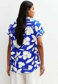 Camicia blu a maniche corte con motivo floreale bianco; vestibilità ampia, orlo arrotondato e texture del tessuto liscia, caratterizzata da un colletto classico.