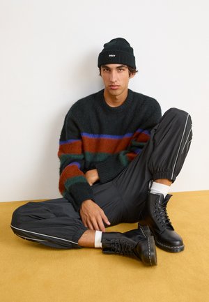 Pull vert foncé surdimensionné avec des rayures multicolores ; pantalon noir à poignets élastiques ; bottes noires à lacets ; assis sur un fond jaune.