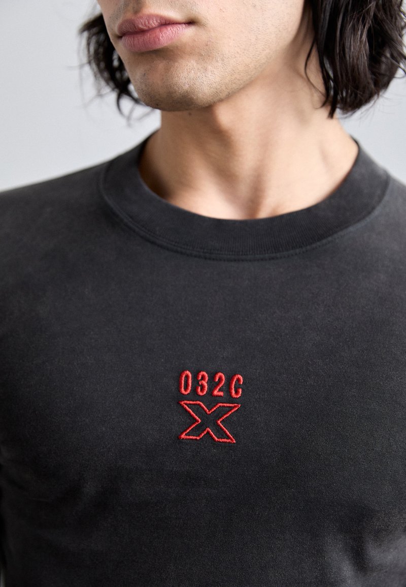 Schwarzer Pullover mit Rundhalsausschnitt, der rot gestickte "032C"- und ein "X"-Logo auf der Brust trägt. Weicher Stoff und minimalistisches Design.