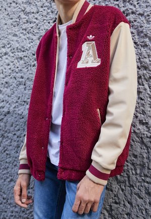 Person trägt eine bordeauxrote Fleece-Collegejacke mit beigen Ärmeln, einem "A"-Logo und blauer Jeans, steht vor einer strukturierten grauen Wand.