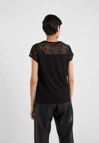 Persona vistiendo una blusa negra de manga corta con encaje en la parte superior de la espalda y los hombros, combinada con pantalones de cuero negros sueltos, de espaldas.