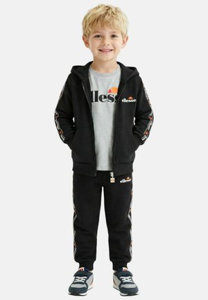Jeune garçon debout, portant un sweat à capuche noir zippé, un pantalon de jogging noir avec des bandes logo, un t-shirt gris et des baskets bleues, souriant à la caméra.