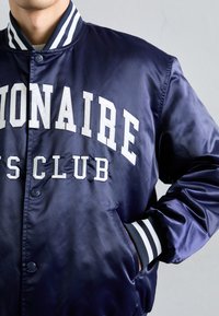 Jachetă bomber din satin navy cu text alb "BILLIONAIRE BOYS CLUB", mansete cu dungi și închidere cu nasture. Include buzunare laterale.