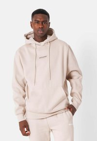 Felpa con cappuccio beige realizzata in tessuto morbido. Presenta una tasca a marsupio, coulisse e un logo stampato sul petto.