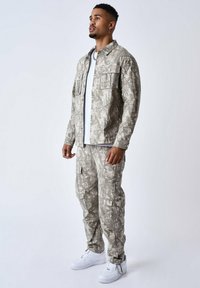 Chaqueta y pantalones cargo con estampado de camuflaje en tonos beige y gris, con grandes bolsillos en el pecho, combinados con zapatillas blancas.