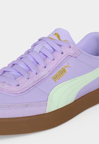 Κοντινό πλάνο ενός παπουτσιού Puma σε λιλά απόχρωση με χρυσό λογότυπο, ριγωτή ταινία σε μέντα, κορδόνια στο ίδιο χρώμα και καφέ λαστιχένια σόλα.