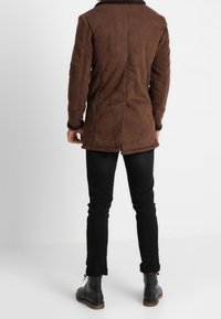 Brun mockapäls med en mörk krage, slim fit, midjehöjd och sidofickor. Bärs över svarta slim-fit jeans och svarta snörkängor.