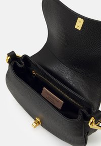 Sac à main en cuir noir avec texture grainée, quincaillerie dorée, fermeture magnétique et poche intérieure zippée. Étiquette de la marque visible à l'intérieur.