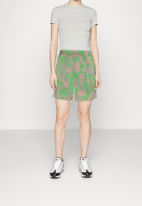 Shorts verts avec un motif de feuilles roses, fabriqués en tissu léger. Associés à un T-shirt gris ajusté et des baskets noir et blanc.